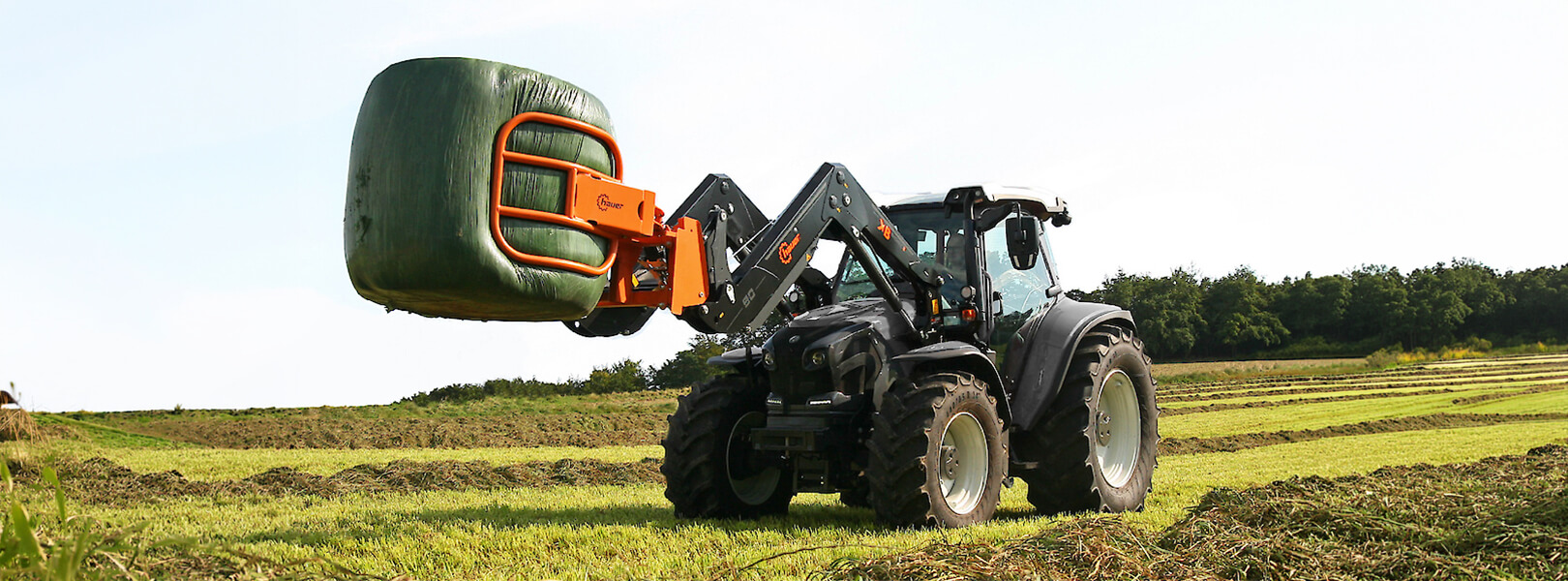 Schwarzer Valtra Traktor mit einem Hauer XB Bionic Frontlader und oranger Ballenzange beim Transport eines Silageballens auf einer Wiese.